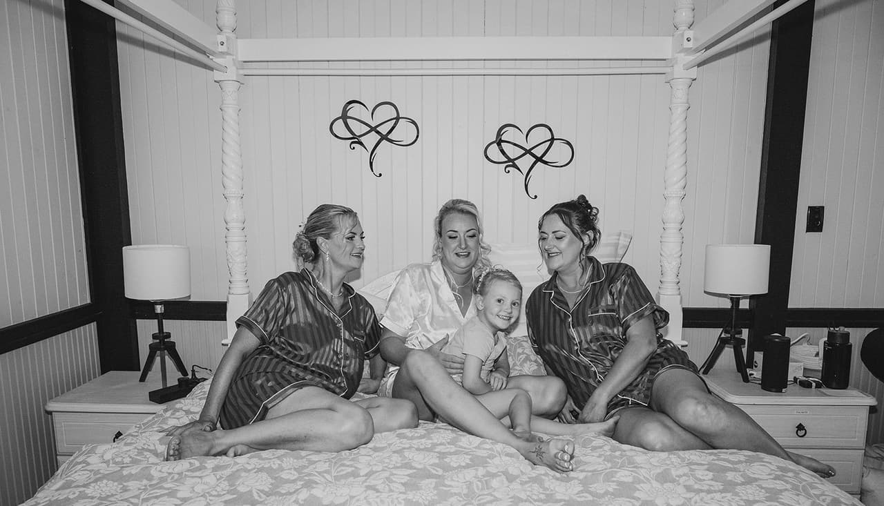 Black and white photo of Jenna and her bridesmaids lying on a four-poster bed with heart decorations above