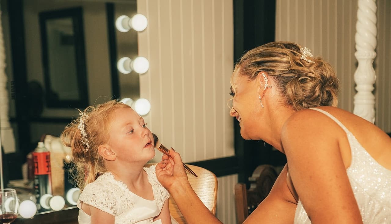 Jenna putting lipstick on a flower girl at the lit mirror