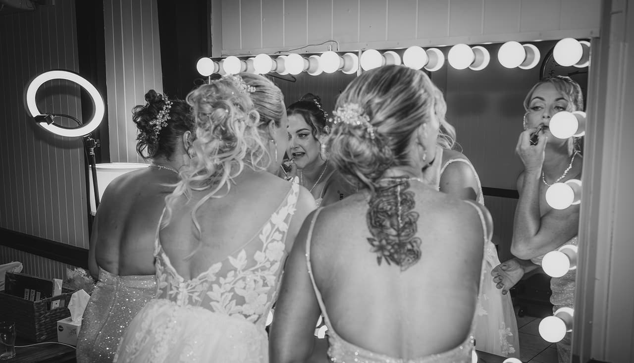 Black and white photo of Jenna and a bridesmaid from behind, reflected in a mirror ringed with bulbs