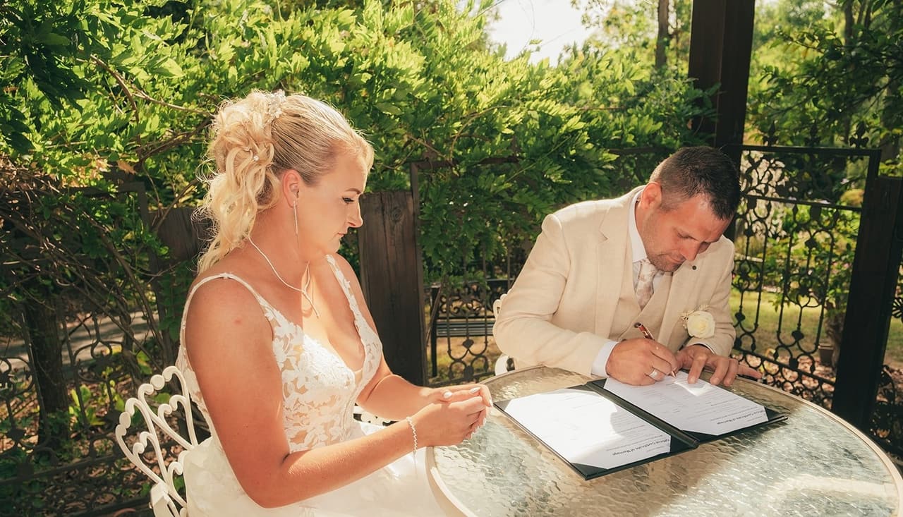 Nick signing the marriage certificate at a glass garden table, Jenna beside him