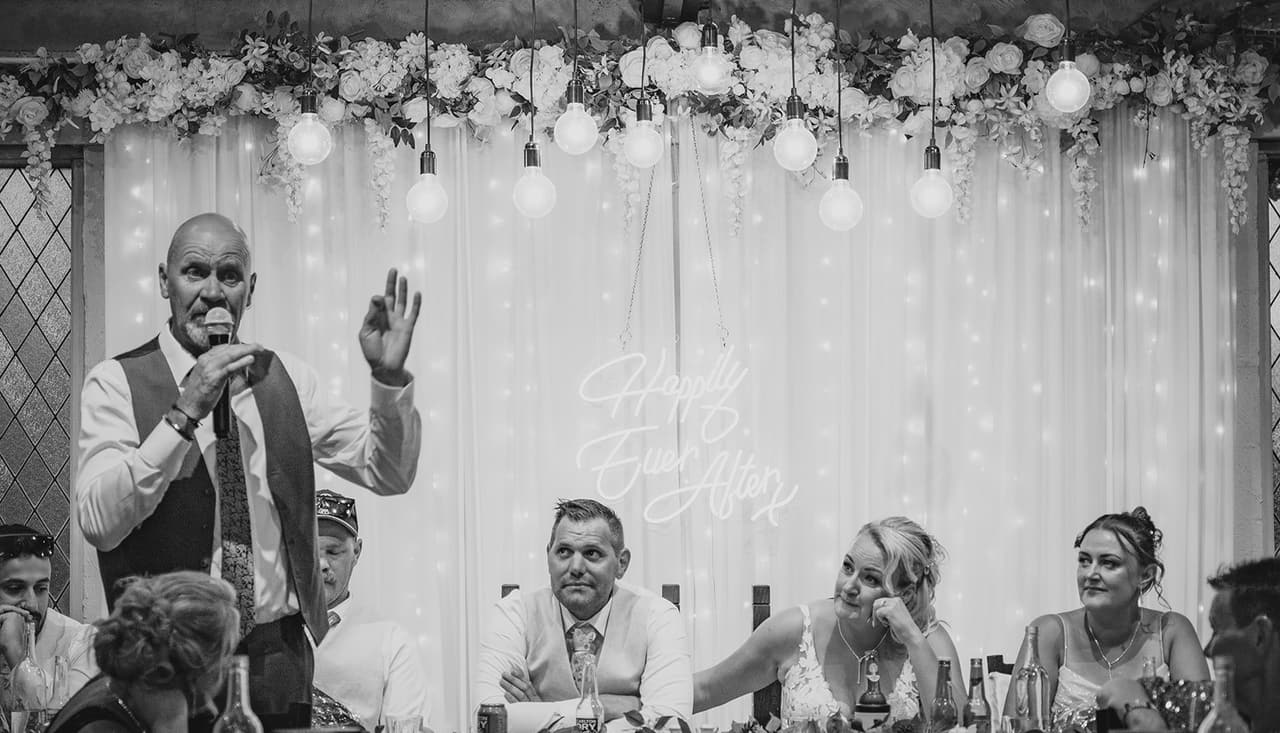 Black and white photo of a speech in front of the curtain of fairy lights at the bridal table