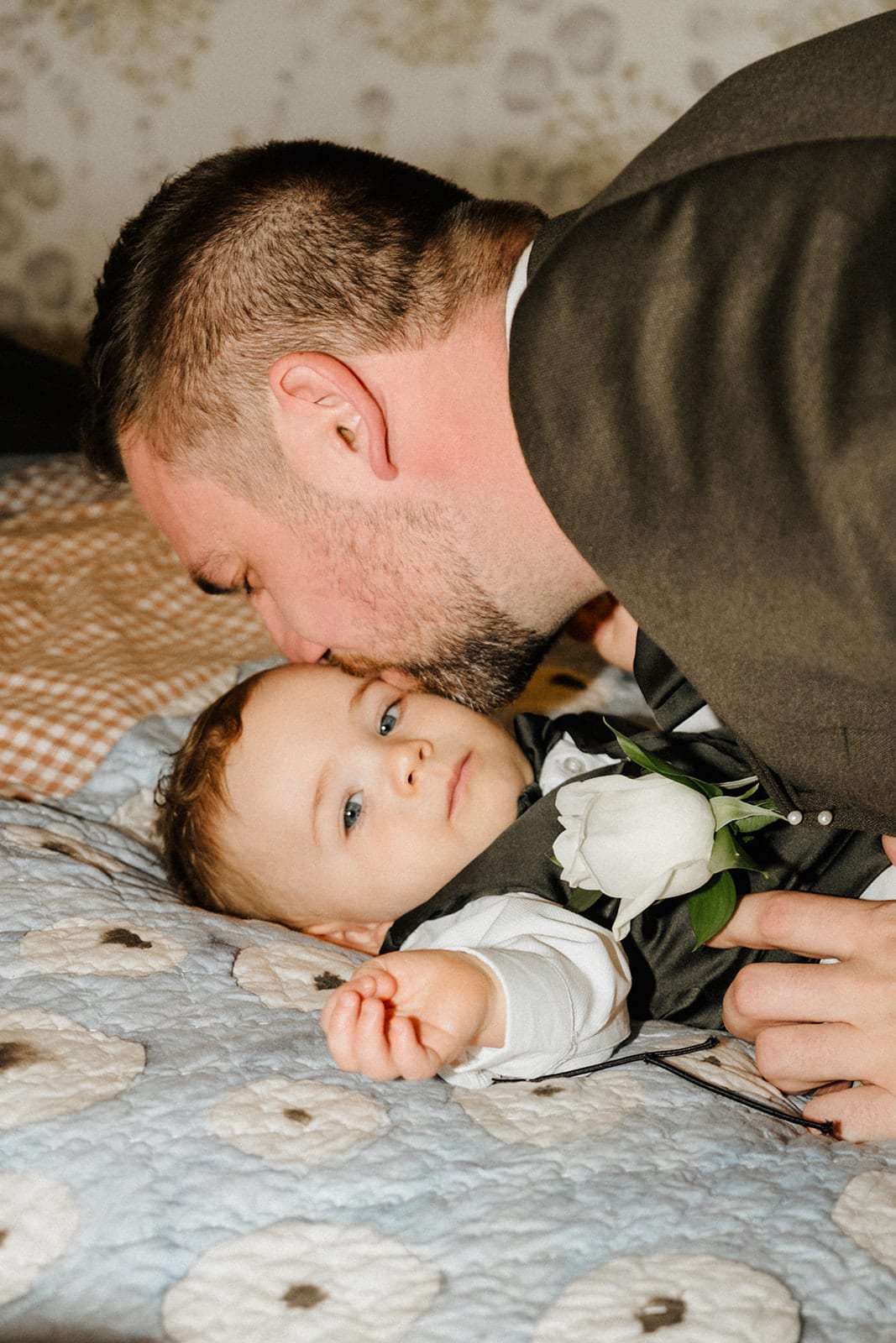 Max, dressed for the day, leaning over to kiss a baby boy lying on the bed