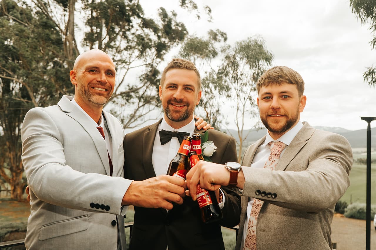 Max and two friends clinking beer bottles on the balcony, gum trees and hills behind them