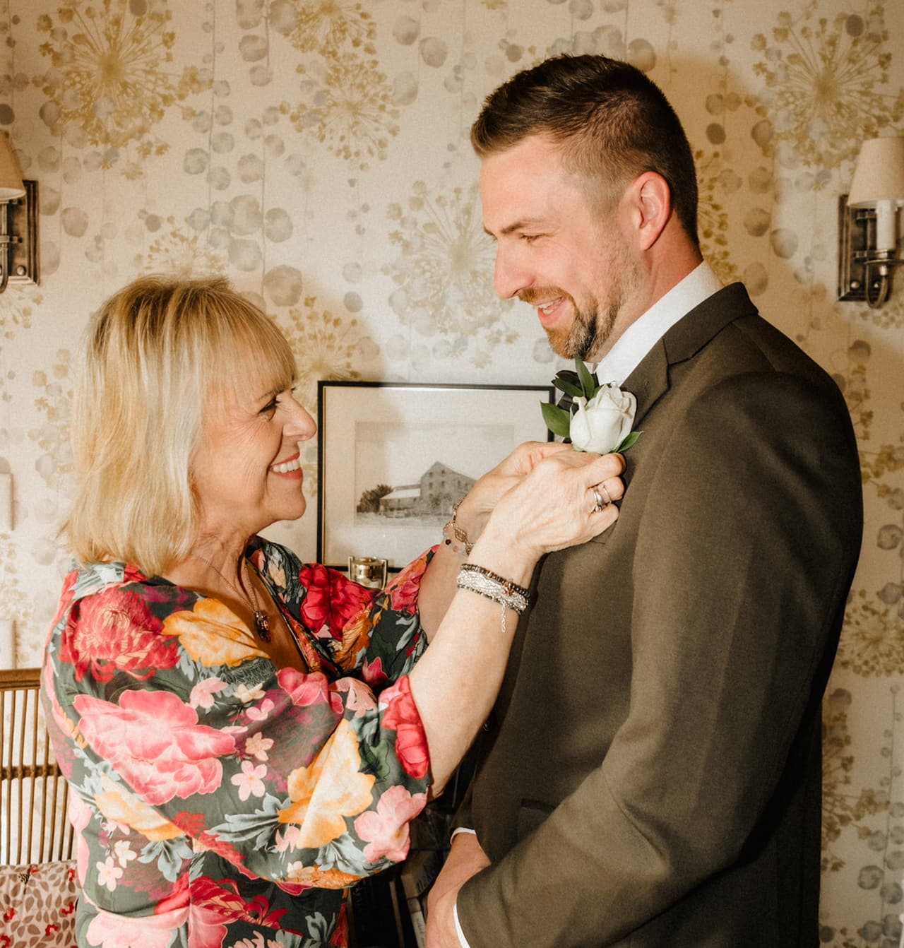 A woman in a red floral dress pinning a white rose buttonhole onto Max’s jacket