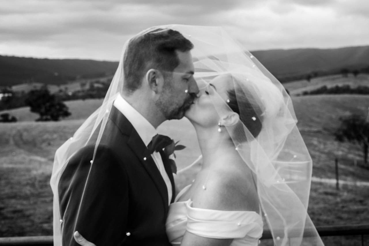 Black and white photo of Jessica and Max kissing under her veil, rolling hills behind them