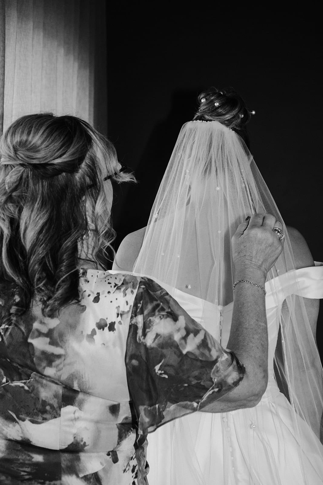 Black and white photo of a woman in a floral top fixing Jessica’s veil into her hair