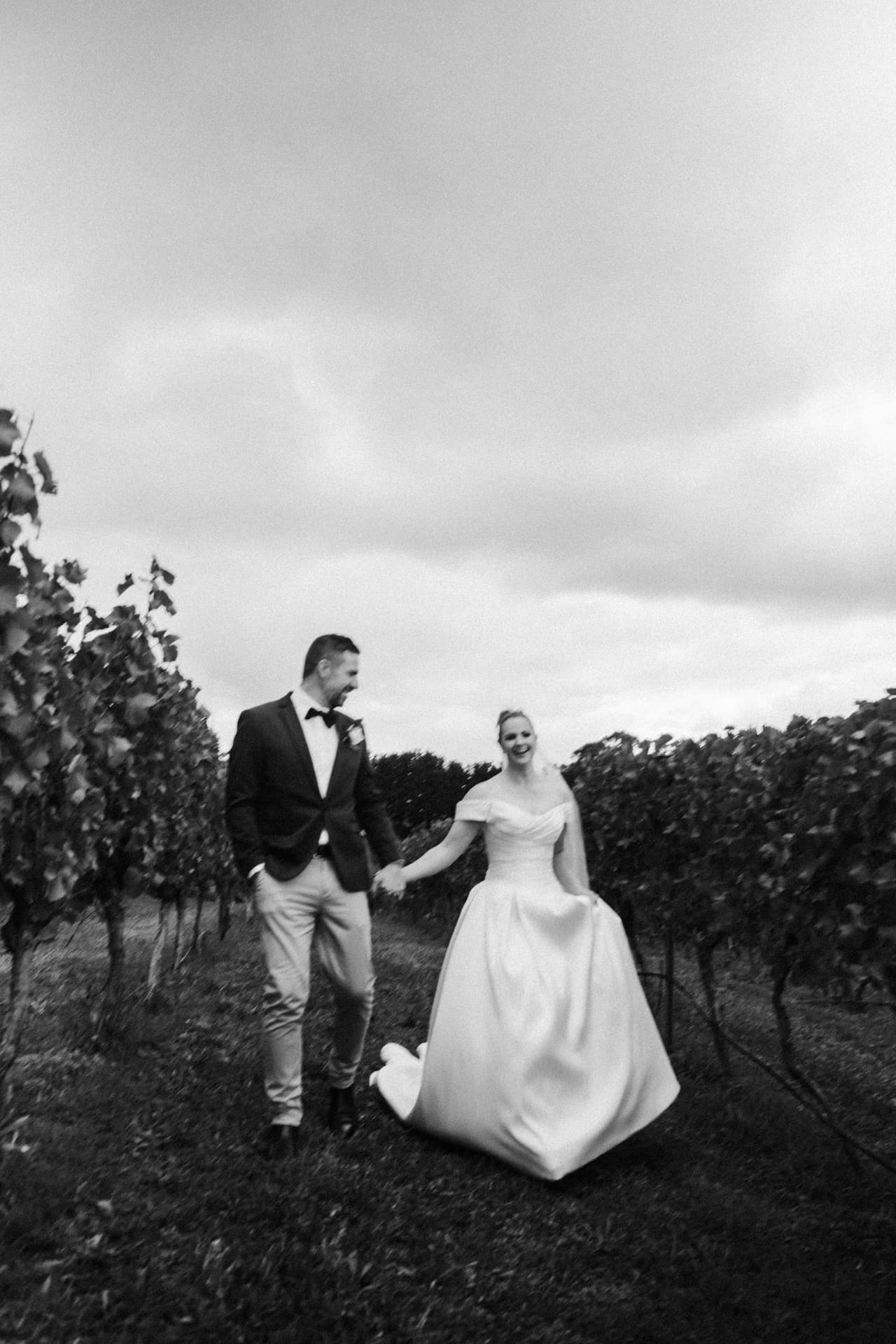 Black and white photo of Jessica and Max walking hand in hand between rows of vines