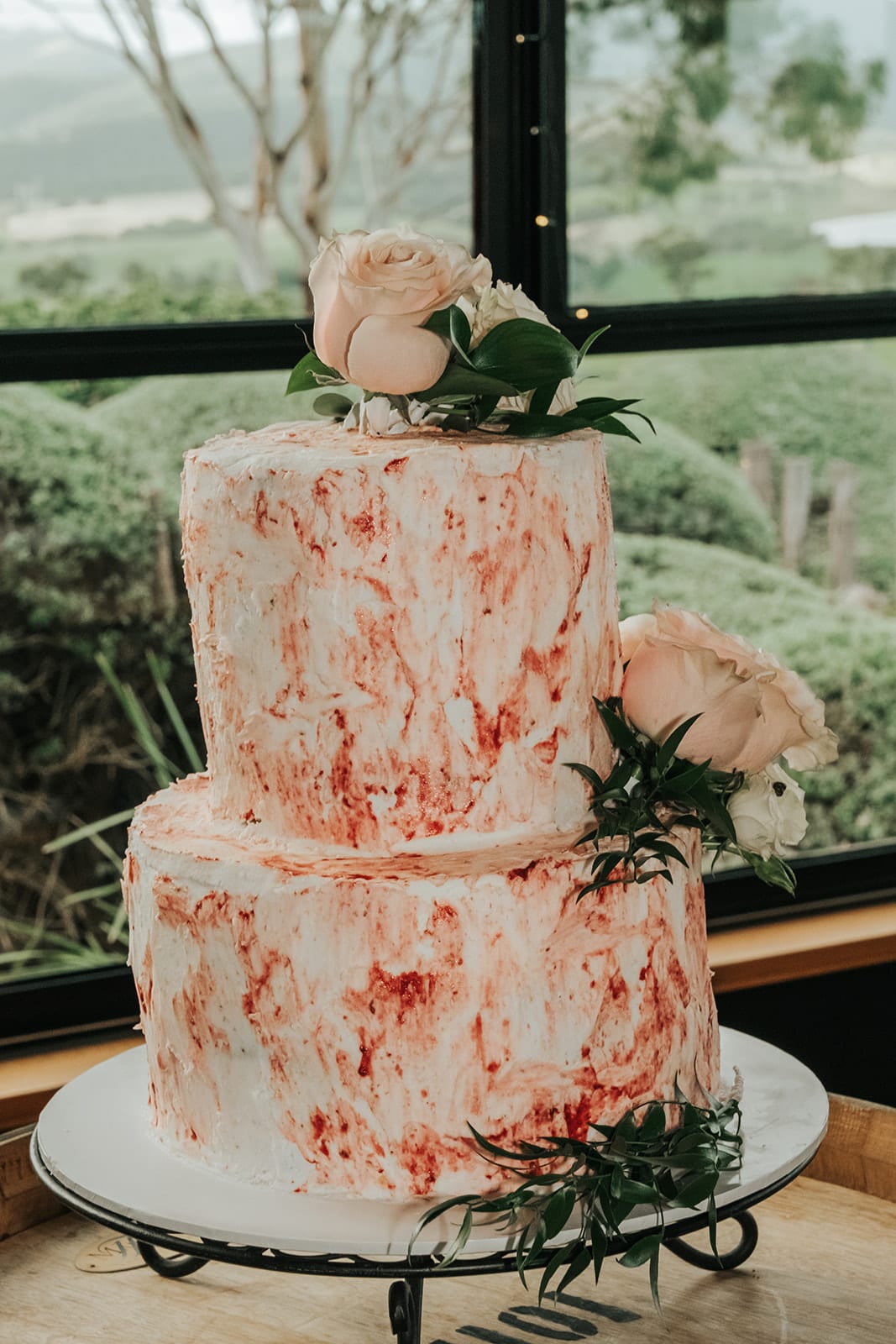 Their two-tier cake with pink marbled icing and roses, standing by a window over the hills
