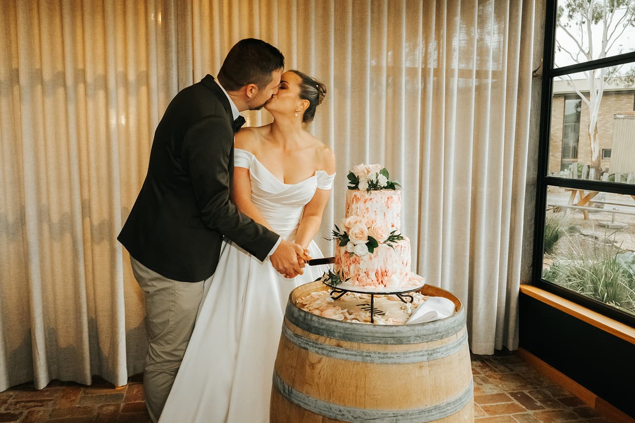 Jessica and Max kissing as they cut their cake on a wine barrel by the window