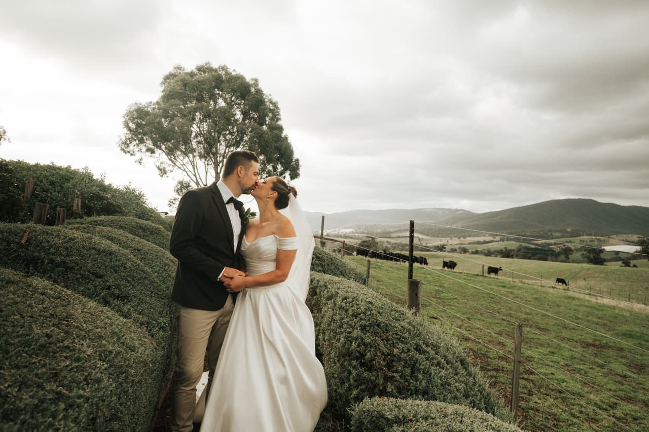 Jessica and Max kissing on a path between rounded hedges, farmland and hills beyond