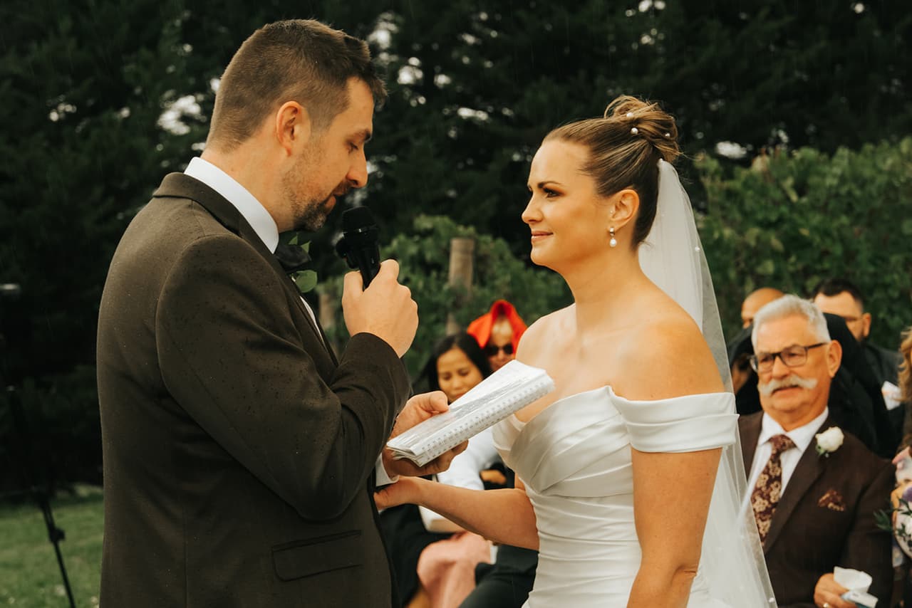 Max reading his vows into a microphone while Jessica listens