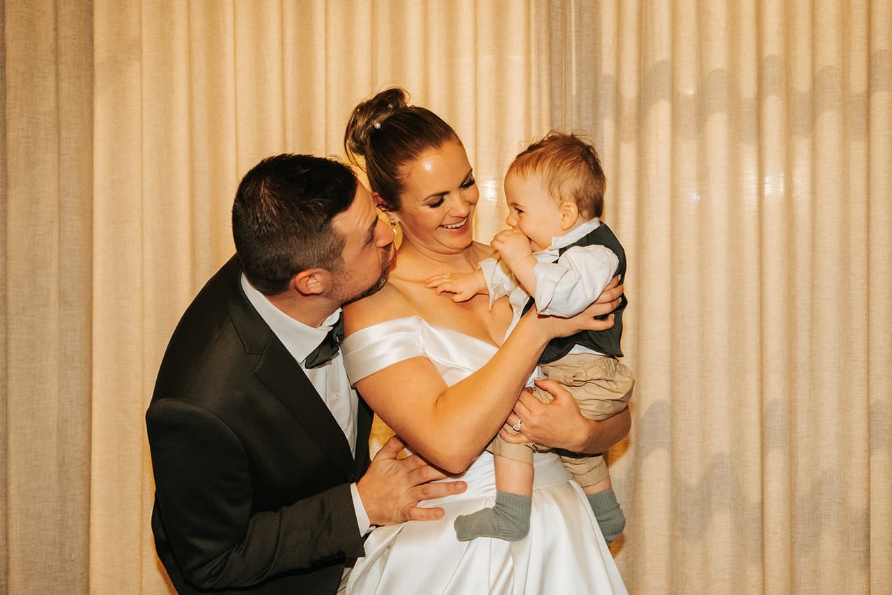 Max kissing Jessica’s cheek as she holds a baby boy in front of a cream curtain