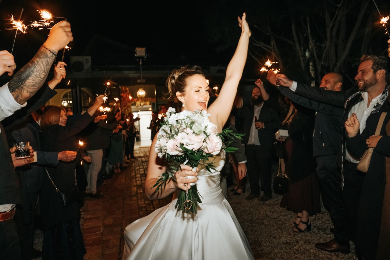 Jessica raising one arm as she and Max walk through a sparkler send-off at night