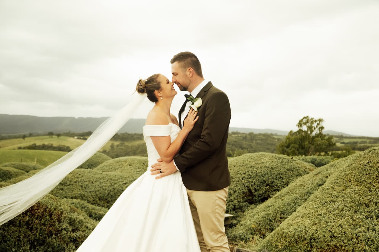 Max kissing Jessica as her veil lifts in the wind over rounded hedges