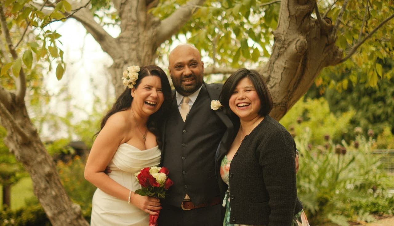 Joanna and Krish laughing under the tree with a guest in a floral skirt