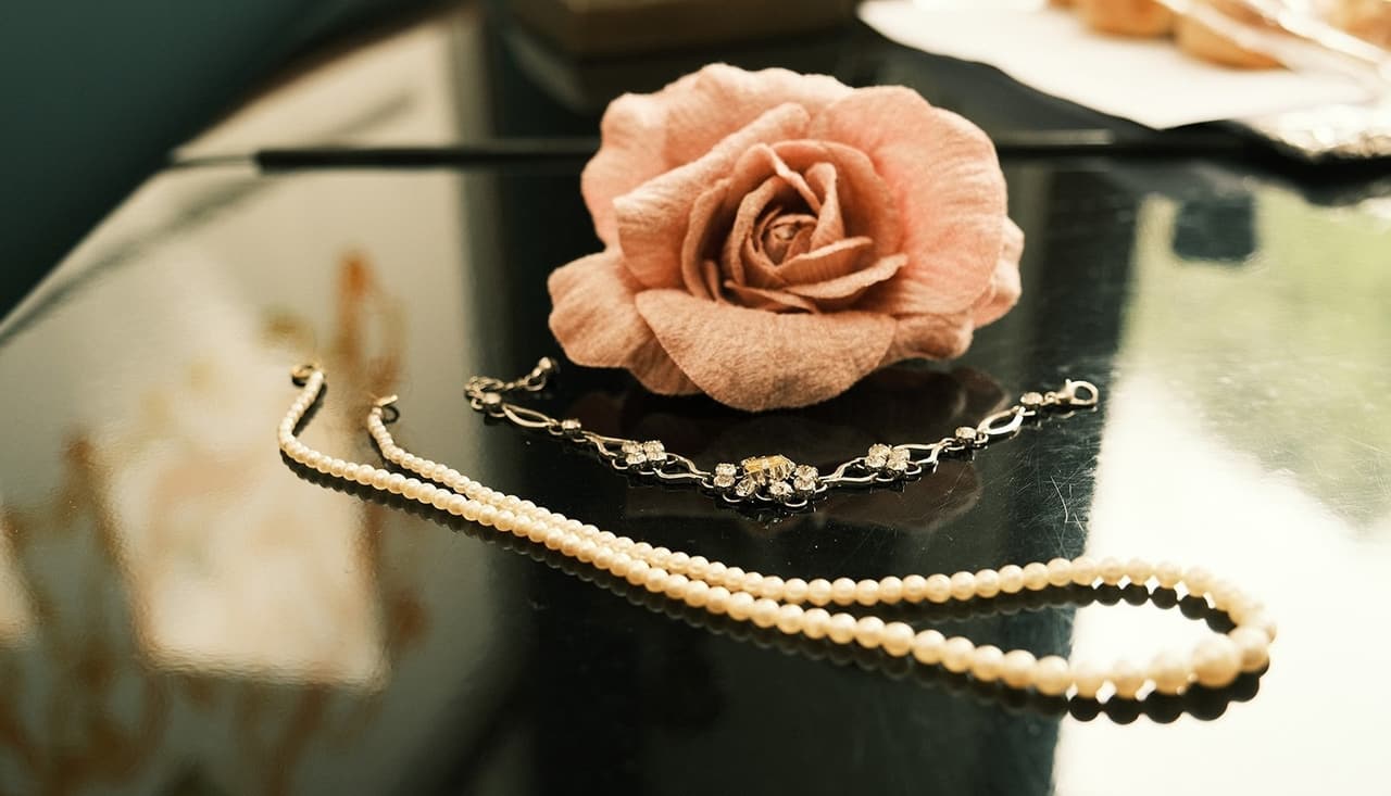 A pink fabric rose beside a string of pearls and a sparkling bracelet on a dark table