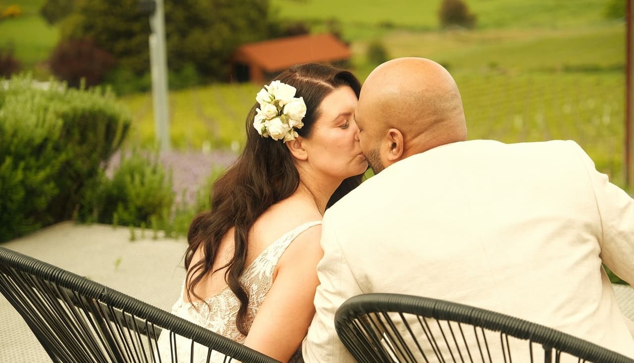 Joanna and Krish kissing between their chairs, the green hills behind them