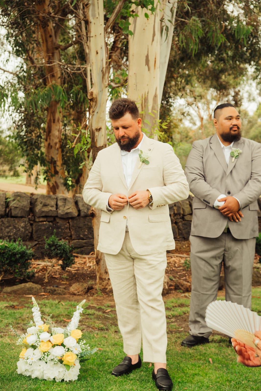Bronson waiting at the front of the ceremony with a groomsman, white and yellow flowers at his feet