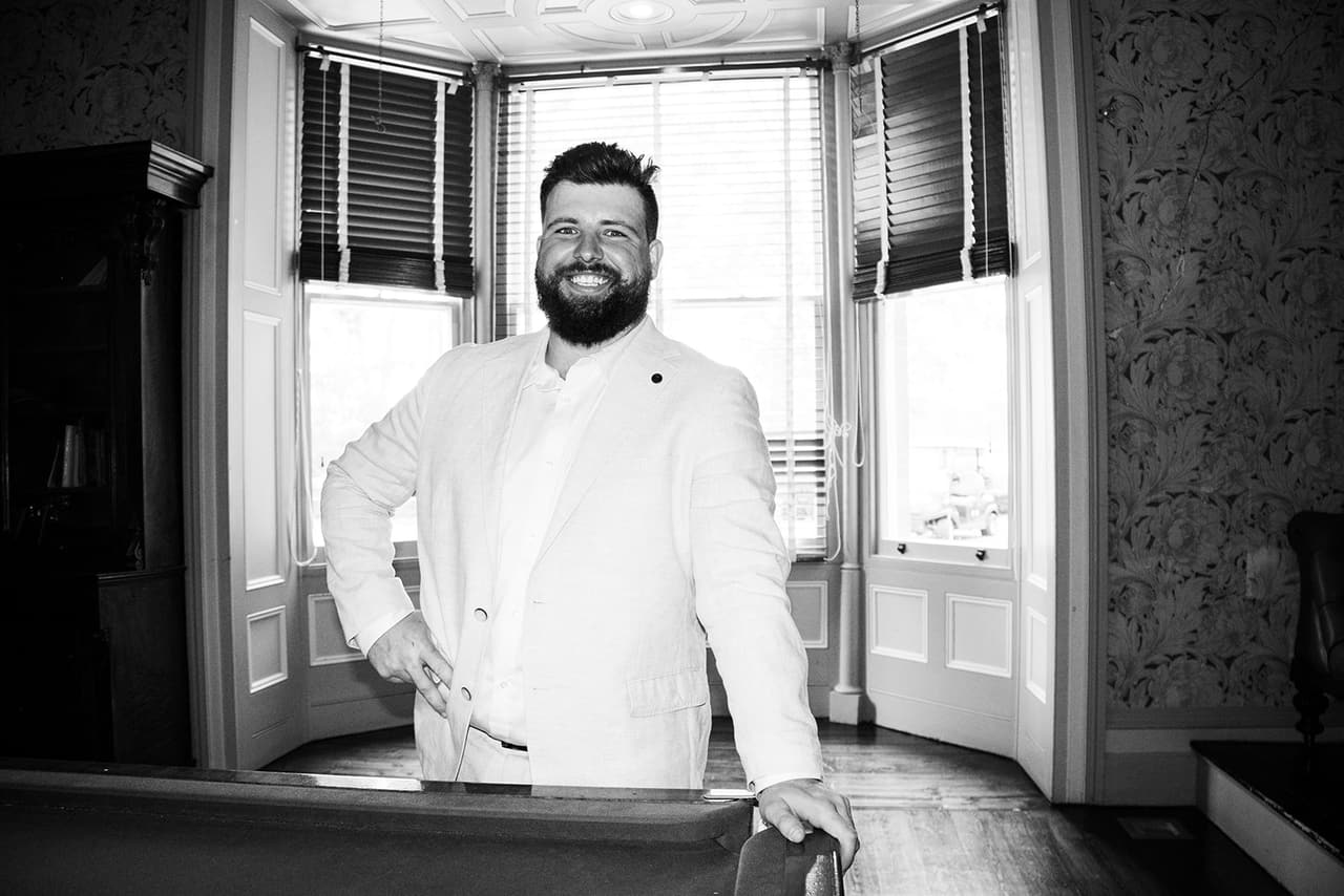 Black and white photo of Bronson smiling in his cream jacket by the bay window