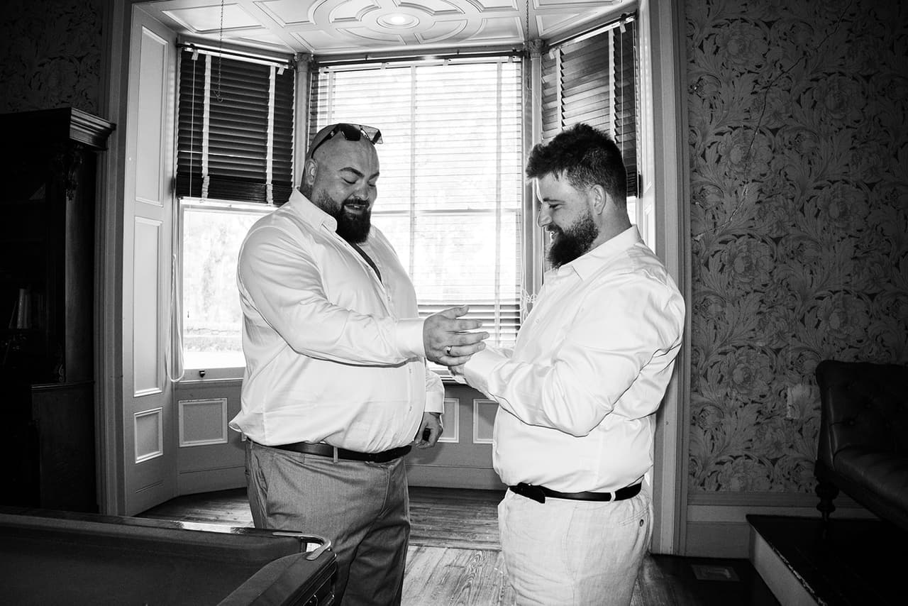 Black and white photo of a groomsman doing up Bronson’s cufflinks in the bay window