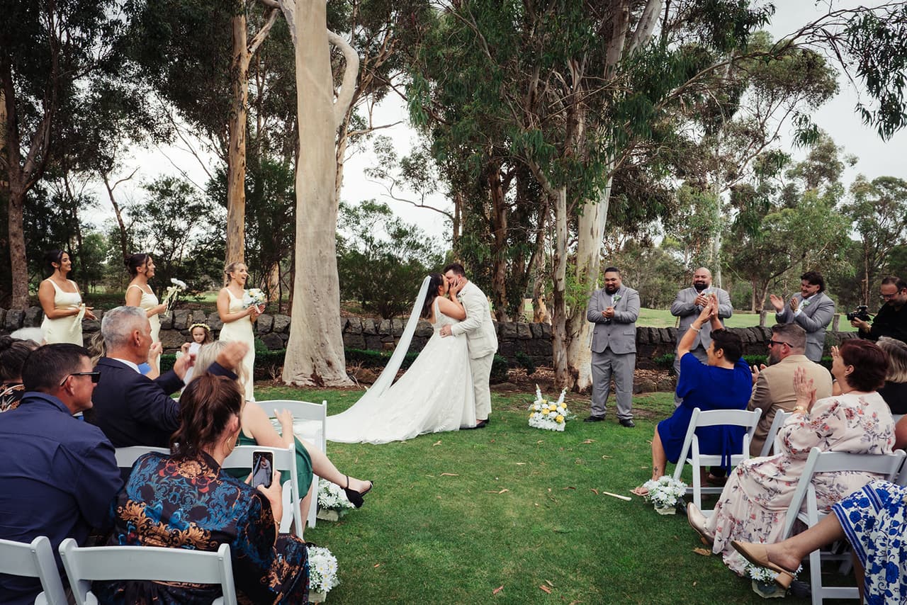 Karina and Bronson’s first kiss under the gums as their guests watch from the white chairs