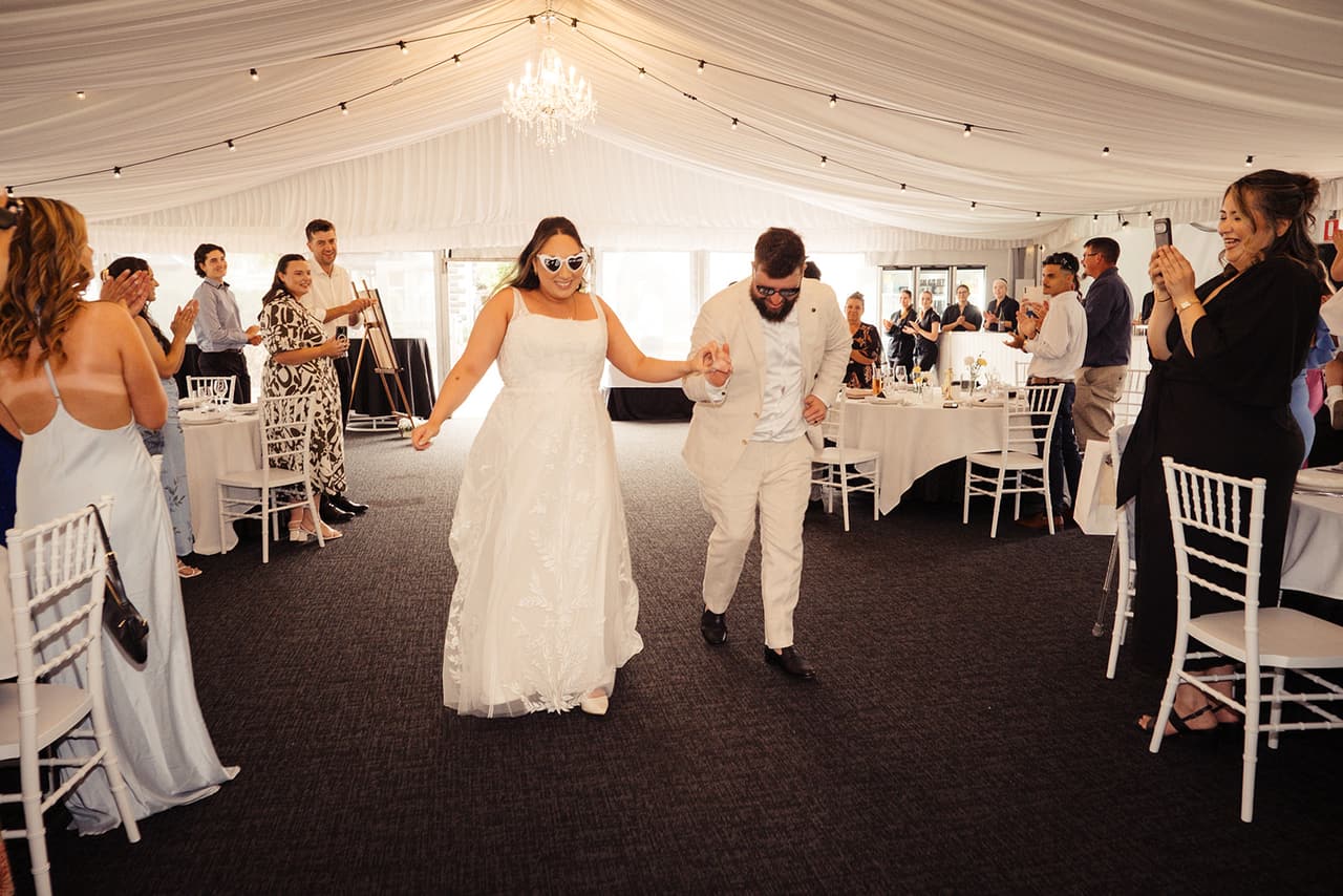 Karina and Bronson walking into the marquee under the chandeliers as their guests stand and cheer