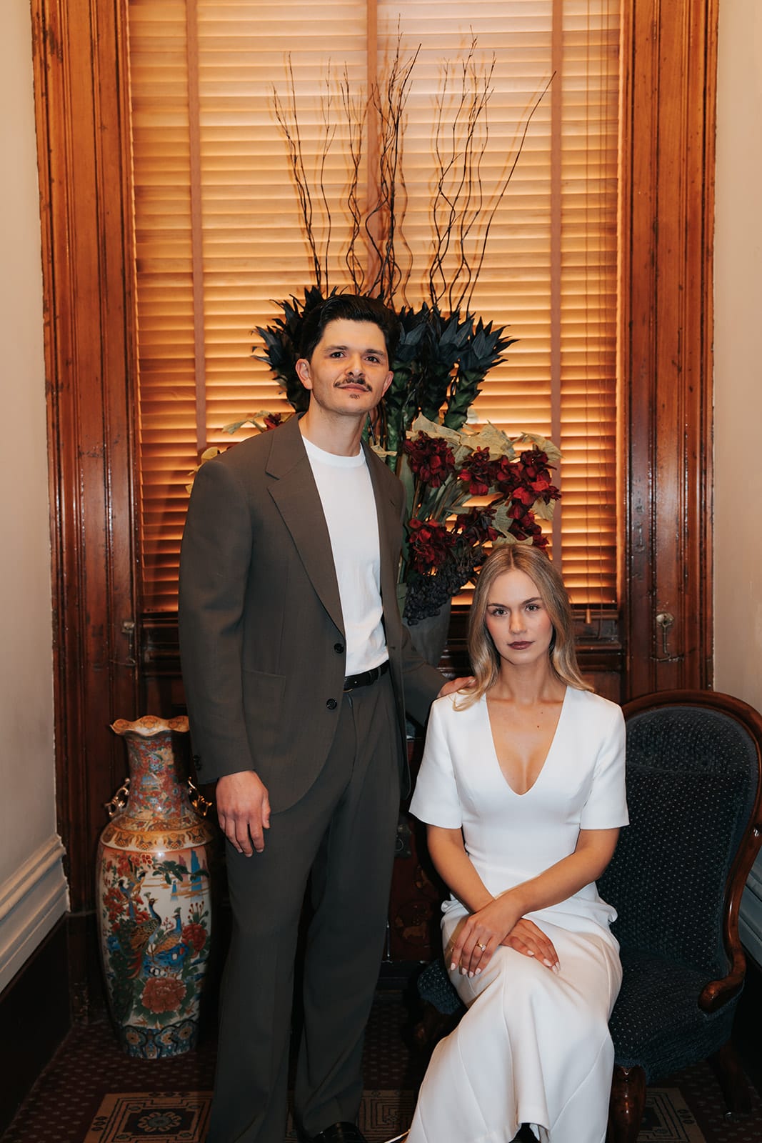 Karina seated in a velvet armchair with Chris standing beside her, a tall vase and timber blinds behind them