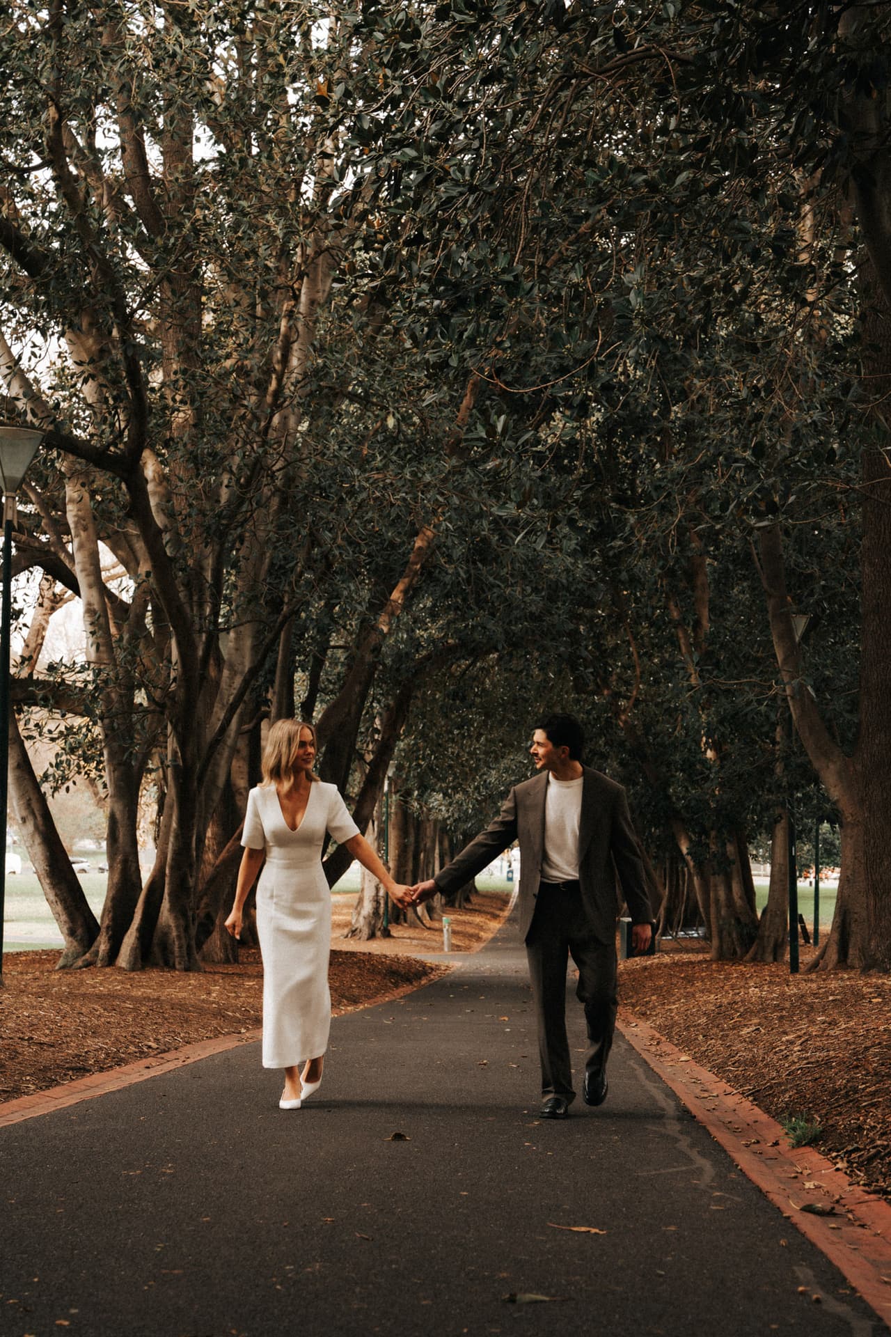 Karina and Chris walking hand in hand along a path under an avenue of old trees