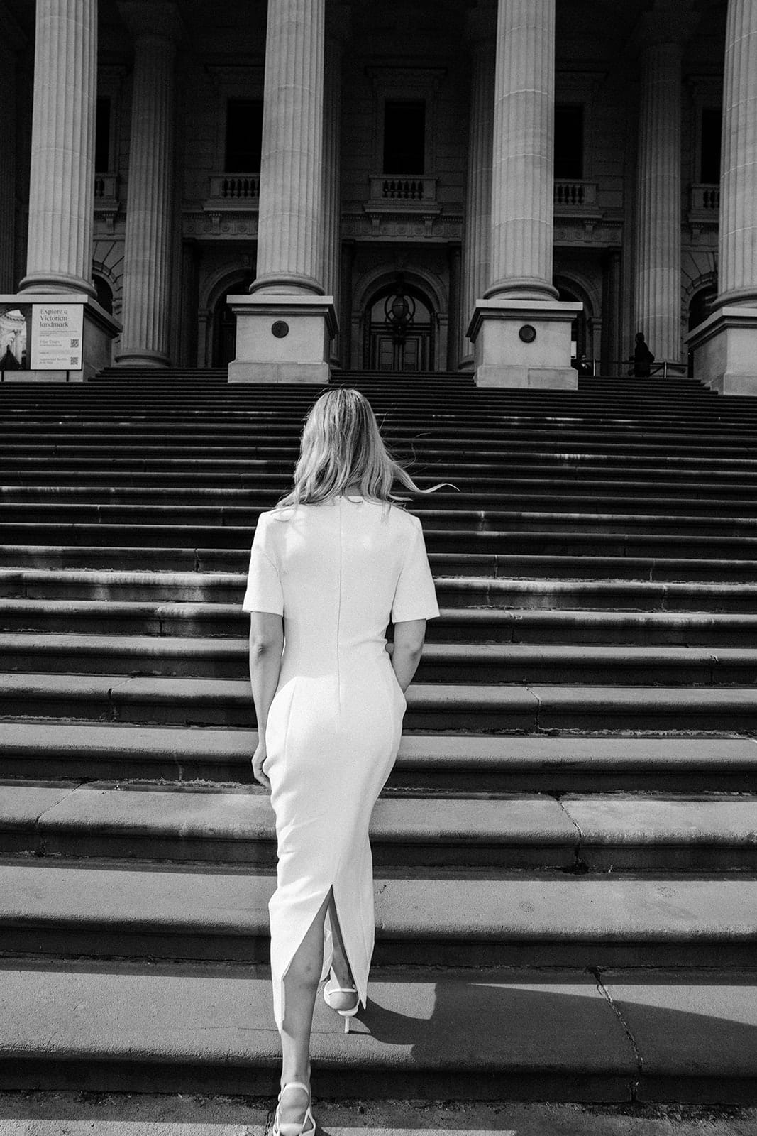 Black and white photo of Karina walking up the wide steps of Parliament House towards the columns