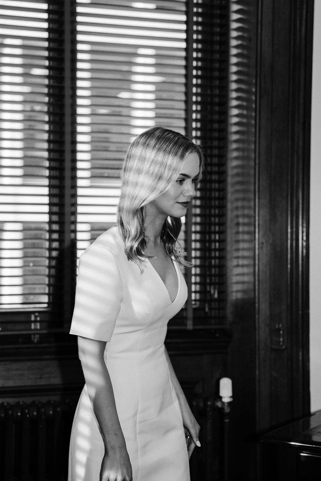 Black and white photo of Karina by a tall shuttered window inside the registry before the ceremony