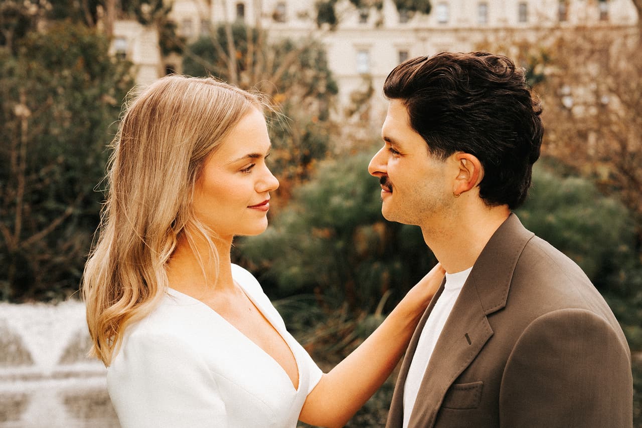 Karina with her hand on Chris’s chest as they look at each other, a fountain playing behind them