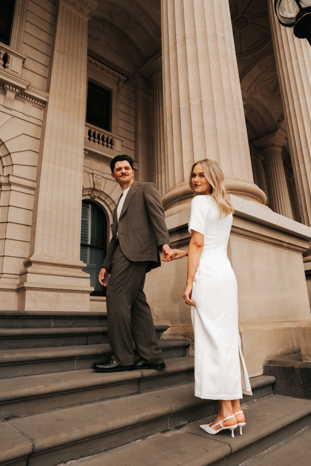 Karina looking back over her shoulder as she and Chris stand on the sandstone steps of Parliament House