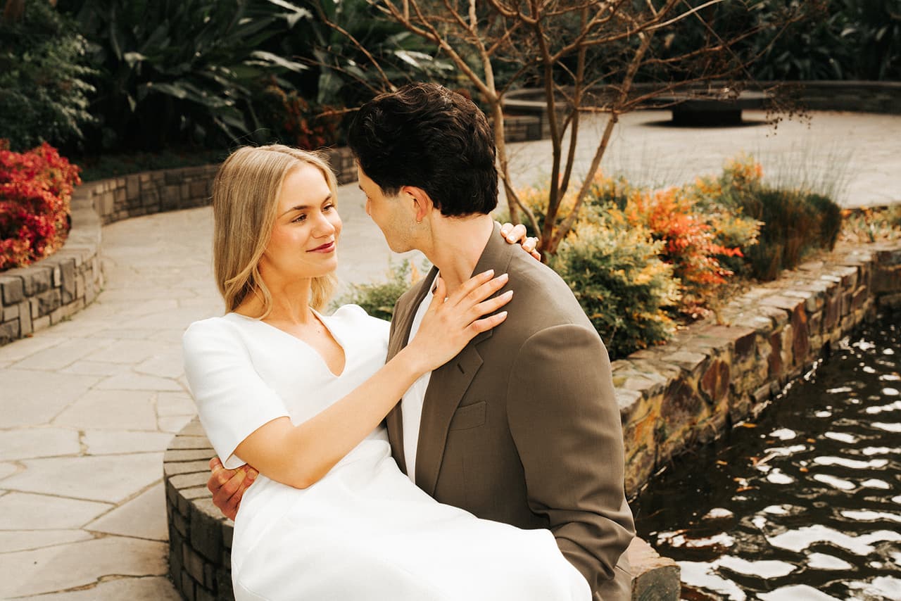 Karina and Chris sitting close on the stone edge of a pond among the garden beds