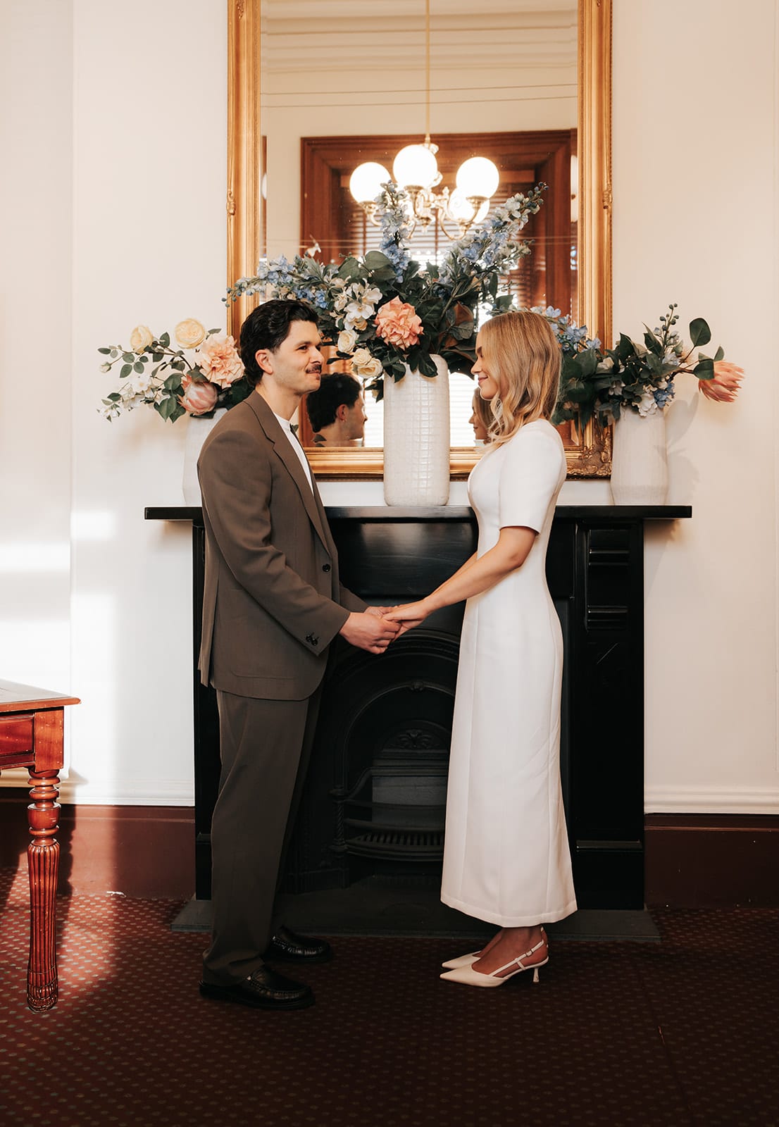 Karina and Chris holding hands in front of the fireplace, a gilded mirror and flowers on the mantel