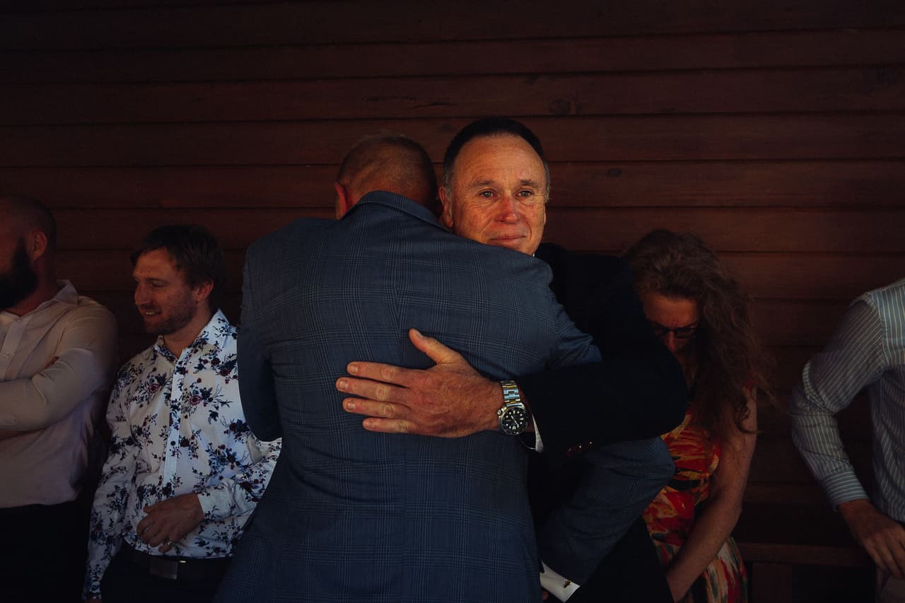 Ben hugging an older guest inside the Boatshed while friends smile around them