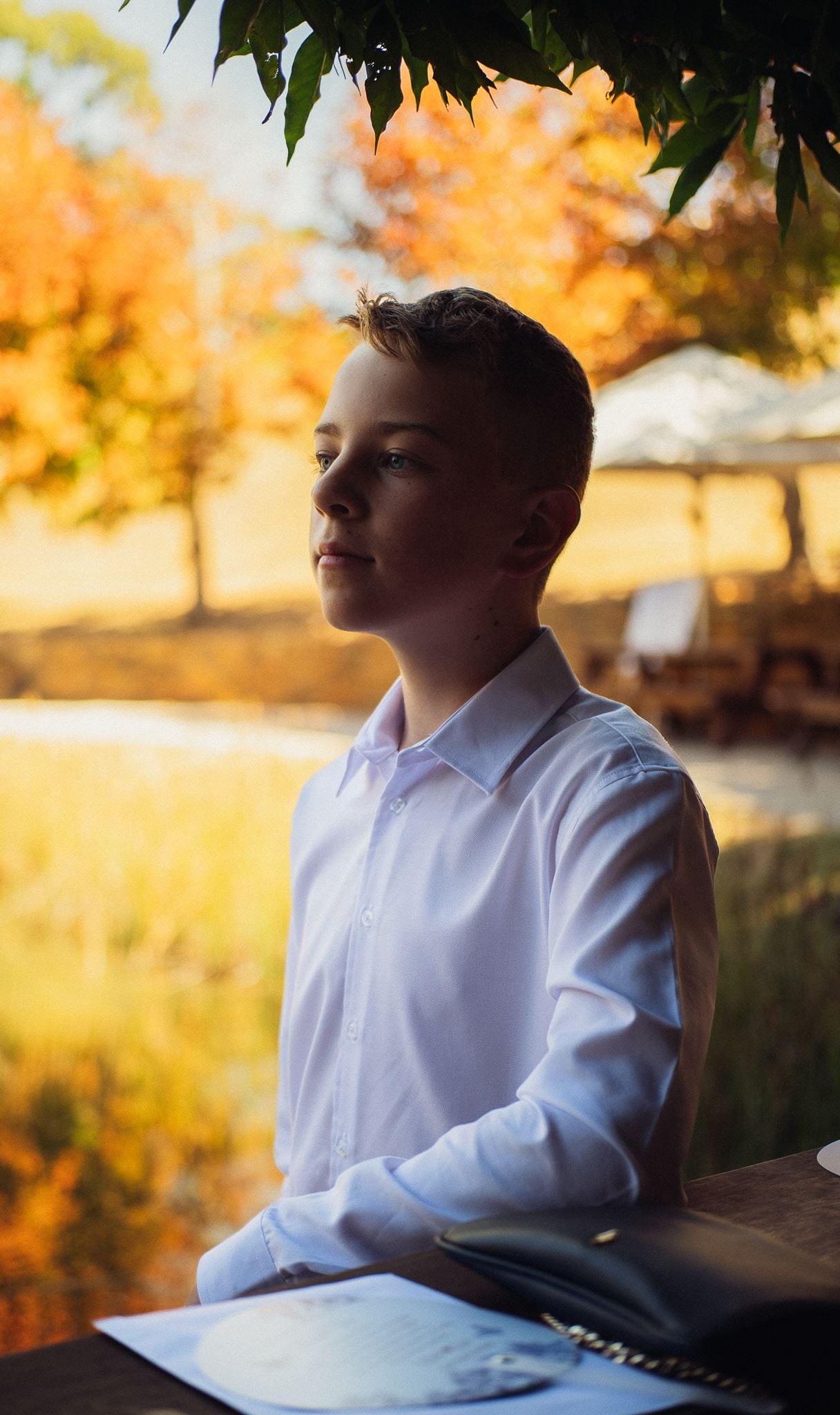 A boy in a white shirt standing at the high table in the Boatshed, autumn trees behind him