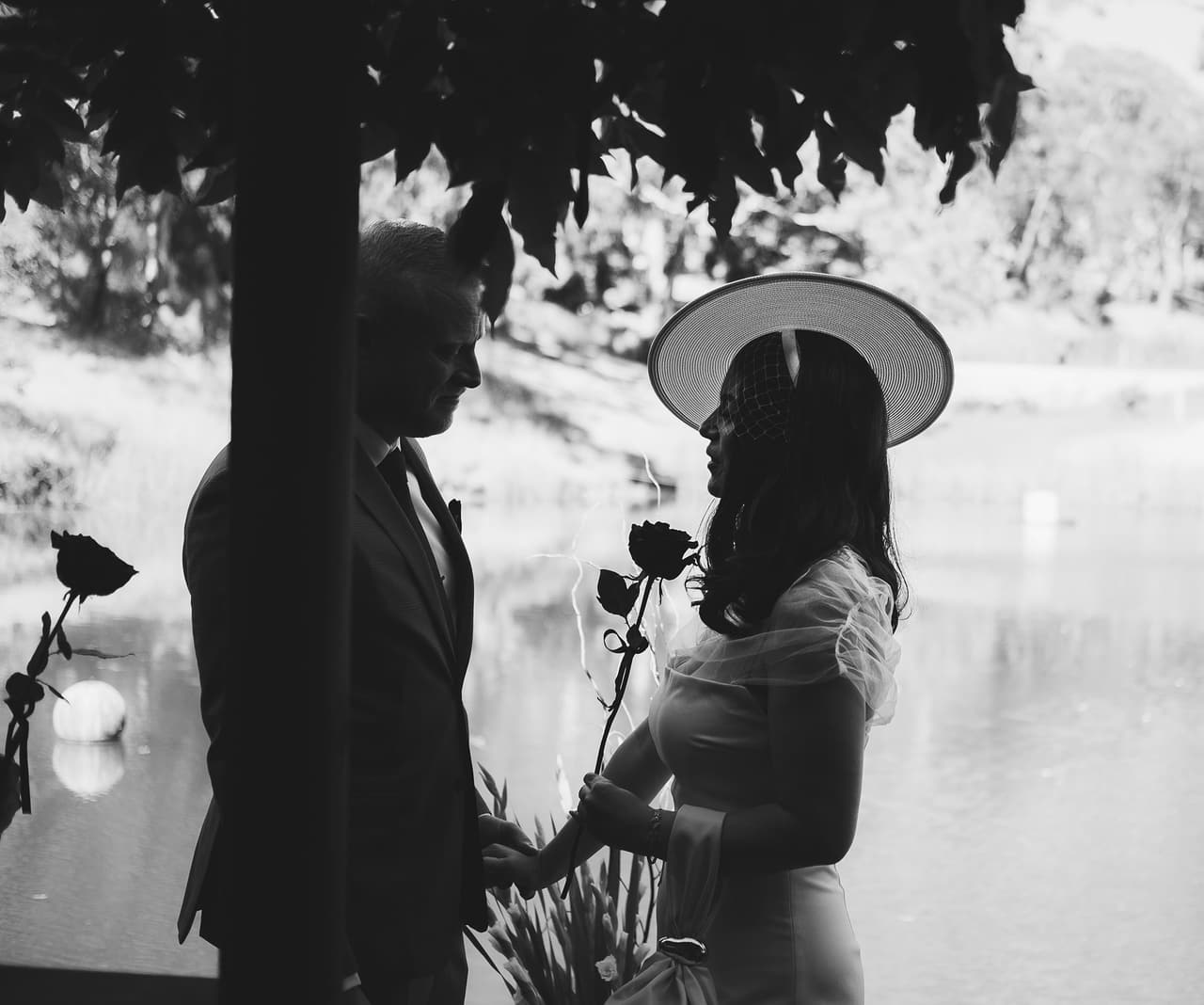 Black and white photo of Kim and Ben holding hands beside a timber post of the Boatshed, Kim in a wide hat with a rose