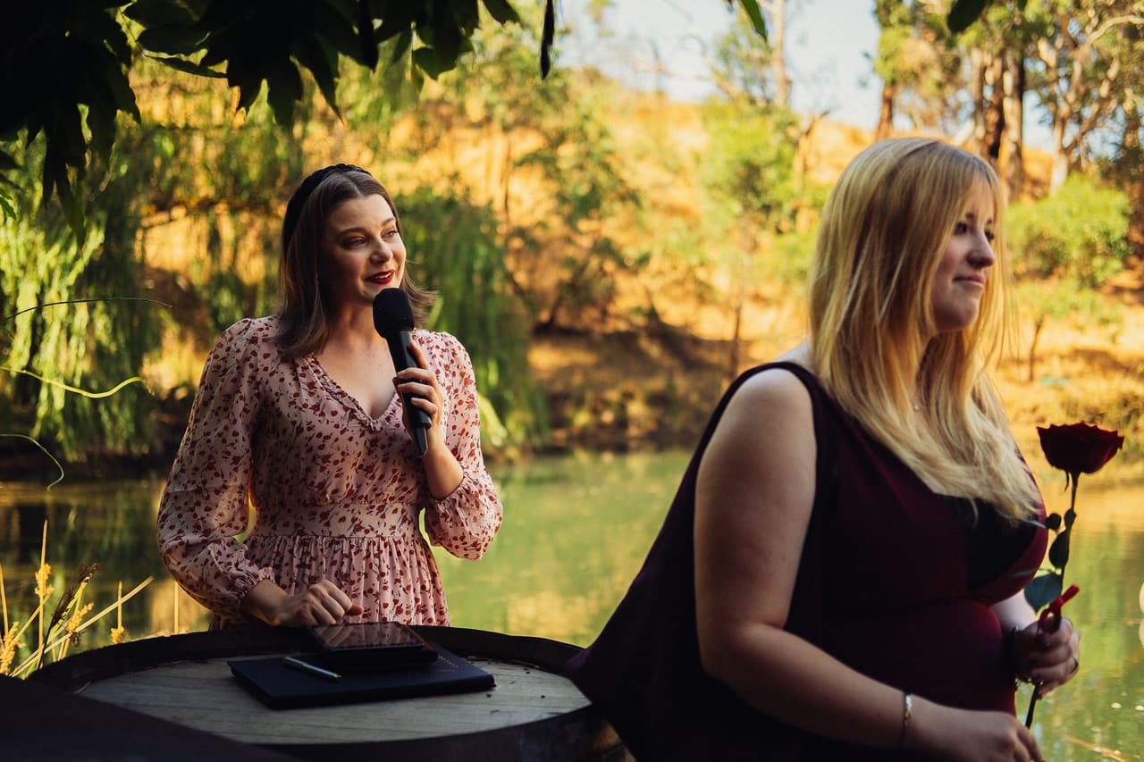 A woman in a floral dress speaking into a microphone at a wine barrel beside a woman holding a red rose