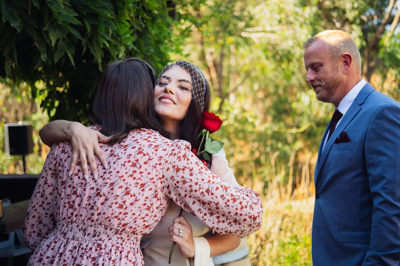 Kim smiling as she hugs a woman in a floral dress, Ben looking on beside the deck