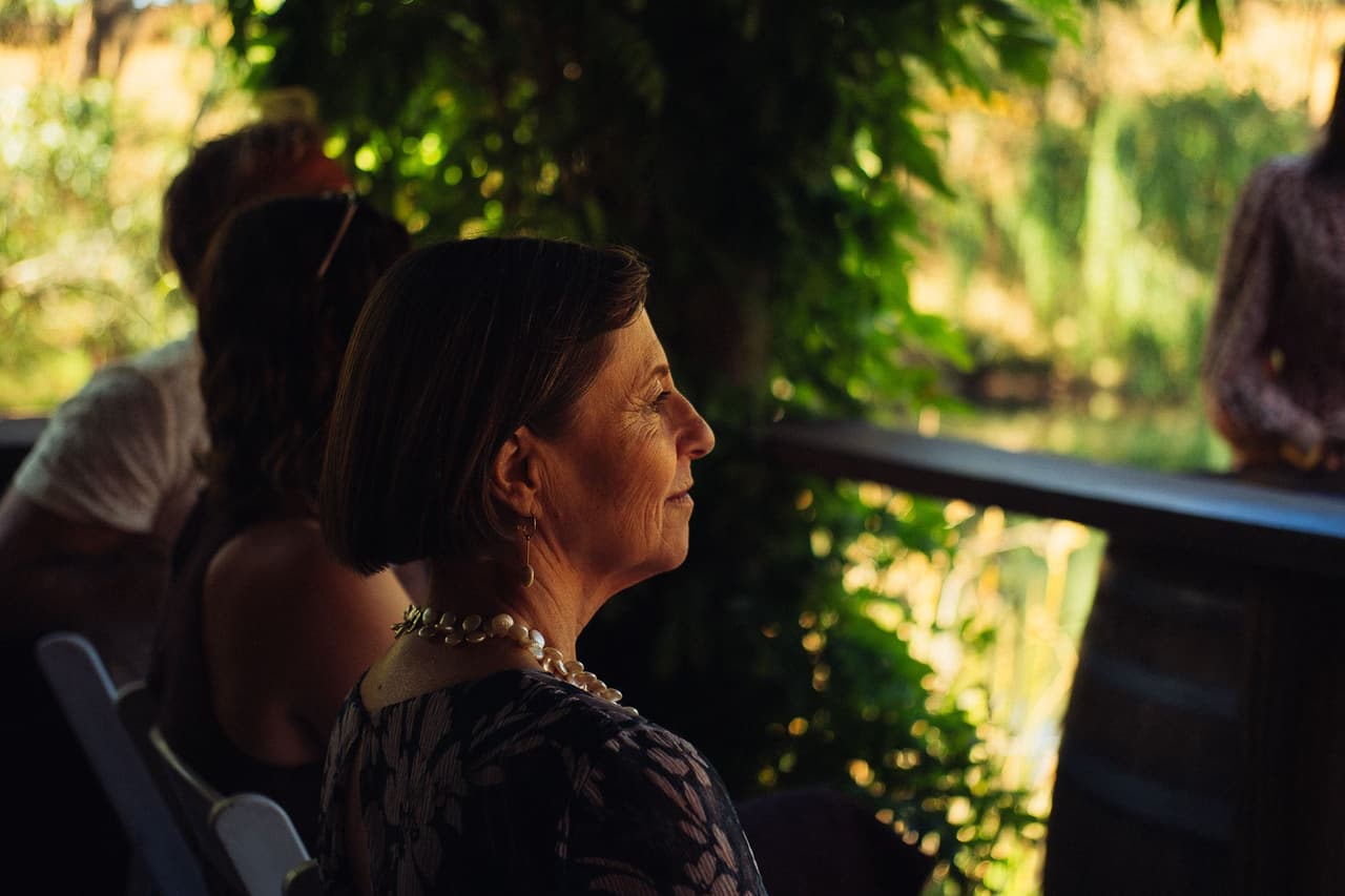An older guest in pearls listening to the ceremony, vines and the pond behind her