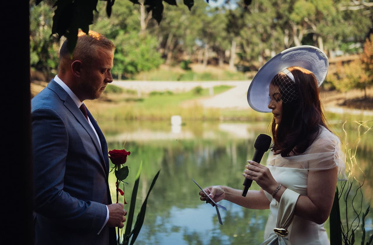 Kim reading her vows into a microphone as Ben listens with a red rose in his hand, the pond behind them