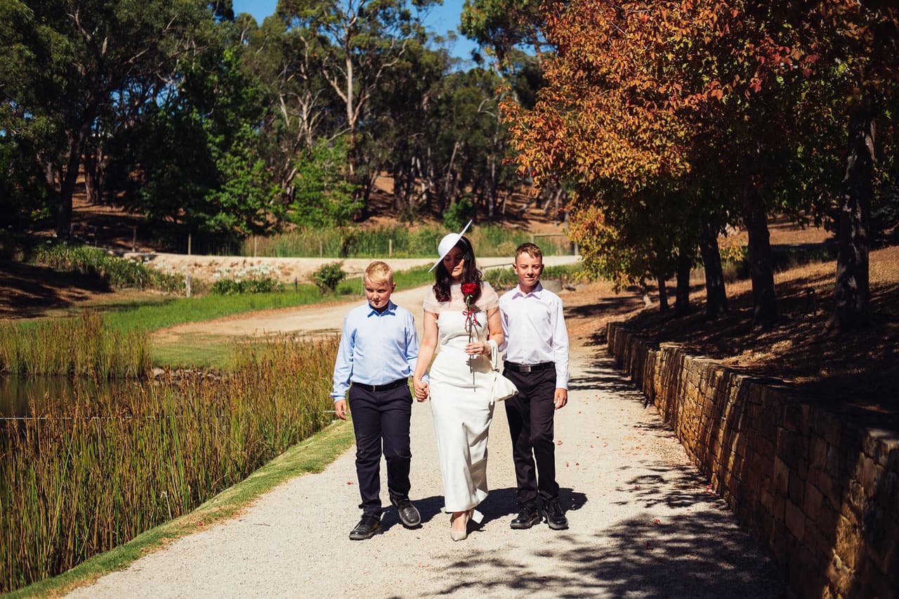 Kim walking along a garden path beside the dam with two boys in blue and white shirts, red-leaved trees on one side