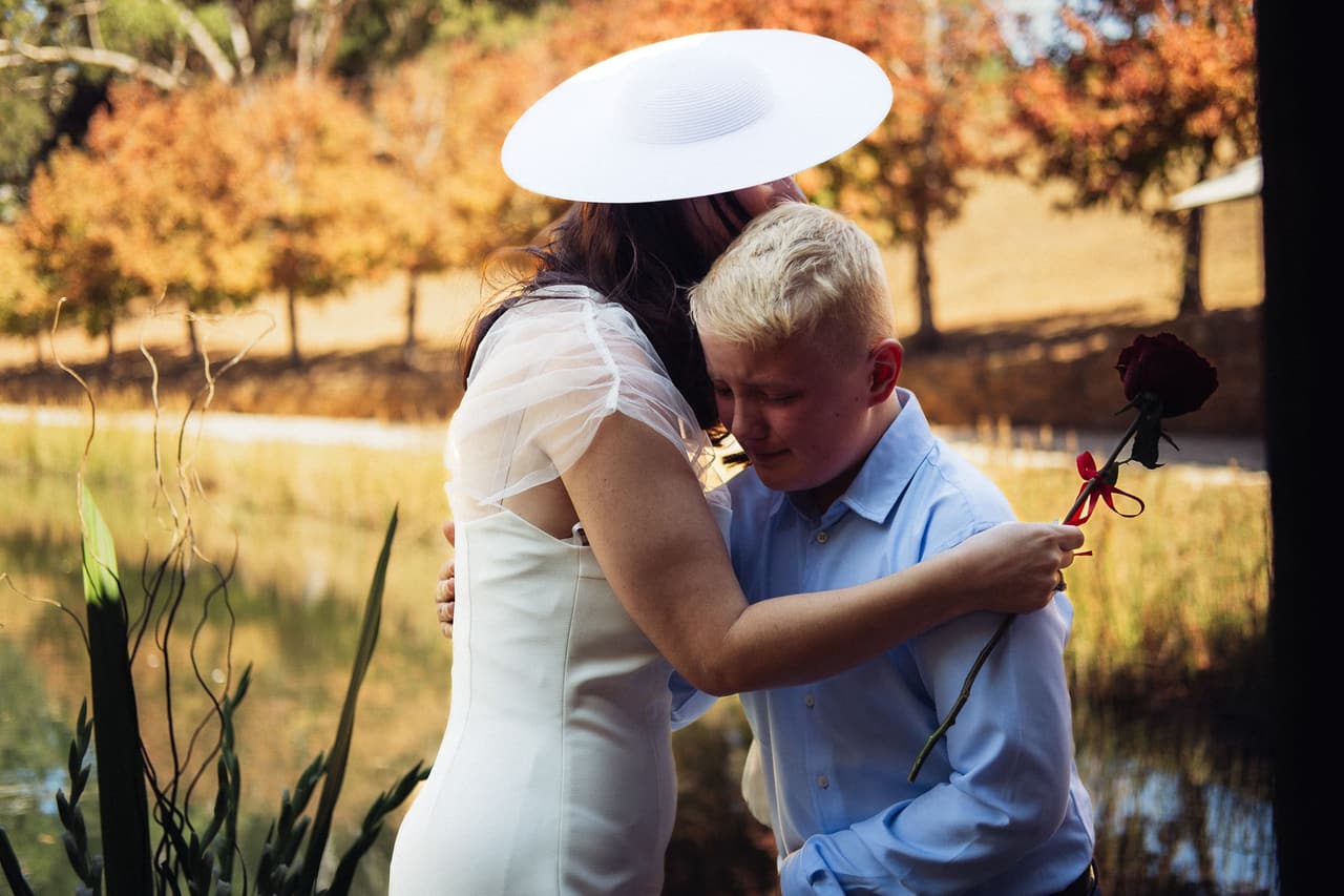 Kim in her wide hat hugging a boy who holds a red rose, autumn trees across the pond