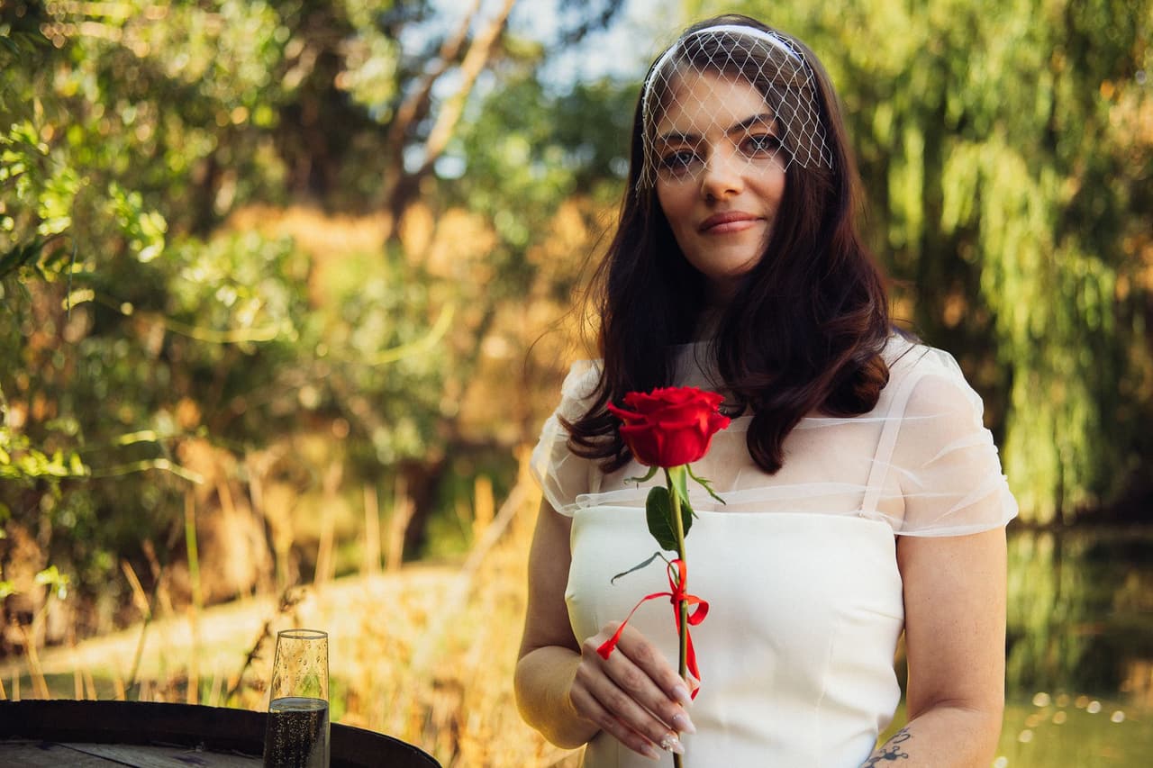 Kim in a birdcage veil holding a red rose tied with a ribbon, the pond and willows behind her