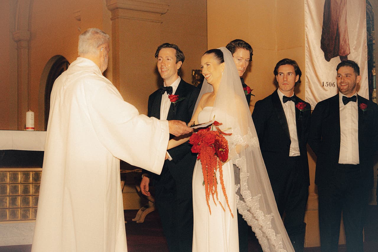 The priest greeting Kira and Nick at the altar as the groomsmen smile behind them