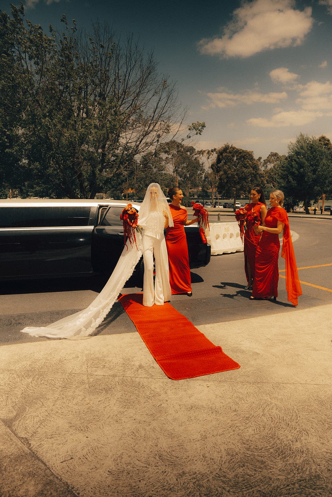 Kira walking along a red carpet from a stretch car outside the chapel, her veil trailing and her bridesmaids in red beside her