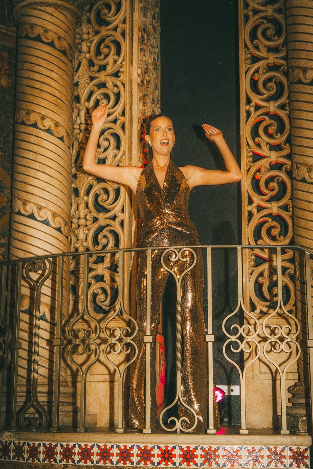 Kira on a Juliet balcony in a gold sequined jumpsuit, arms raised