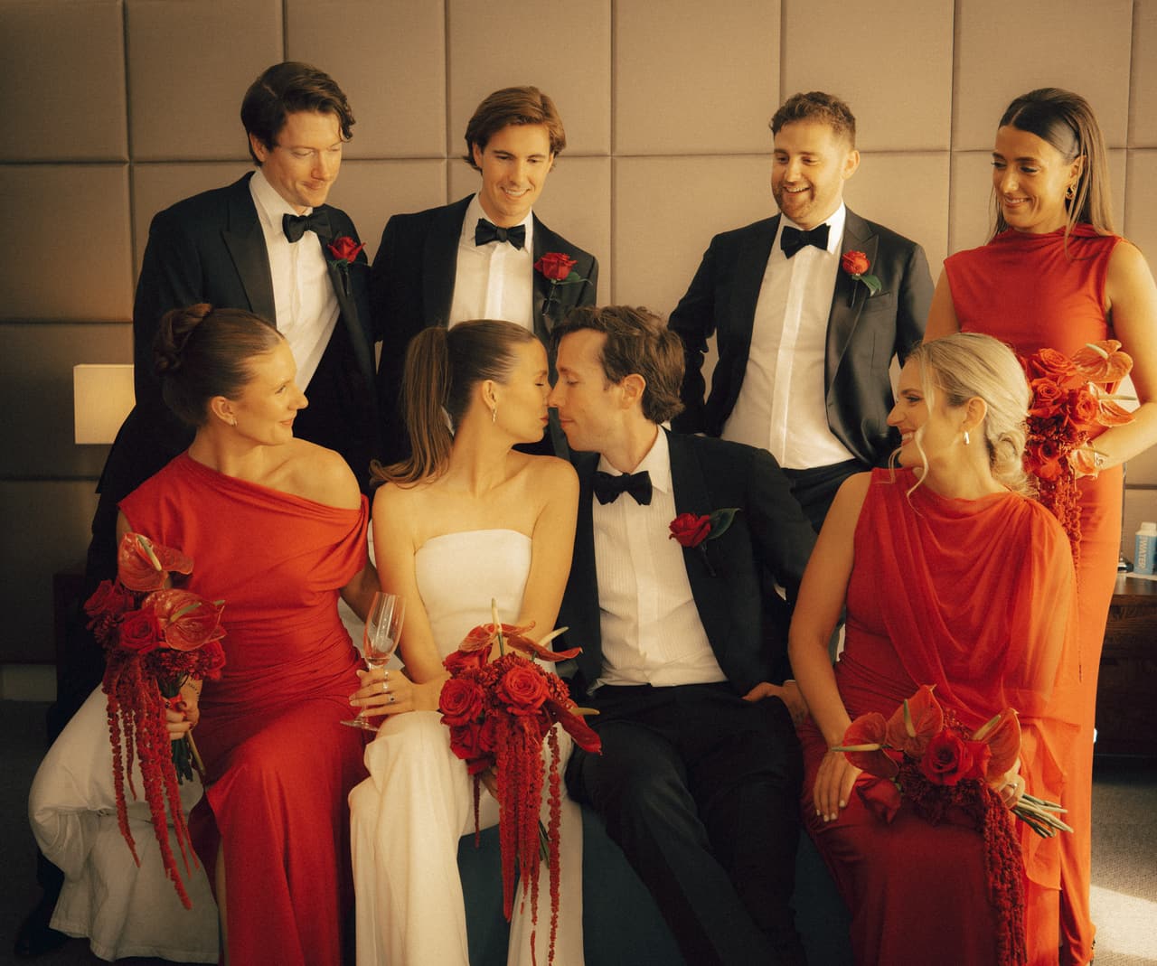 Kira and Nick kissing on a long banquette among bridesmaids in red and groomsmen in black tie