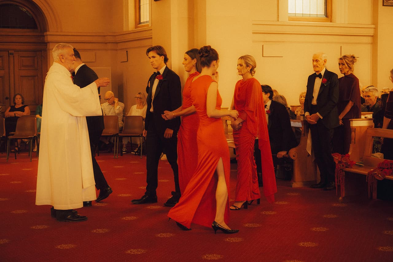 Groomsmen and bridesmaids in red filing past the priest at the front of the chapel during the ceremony
