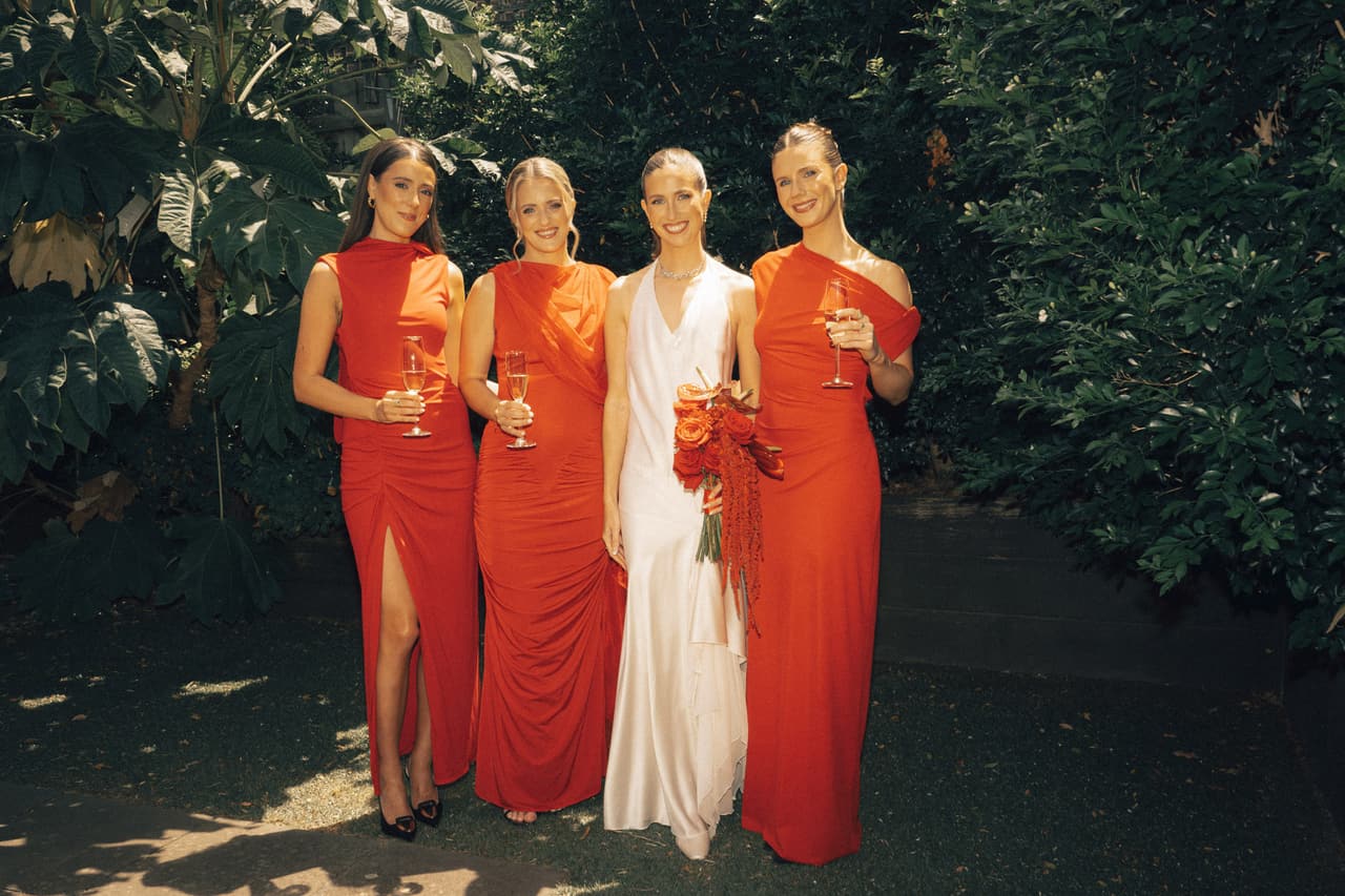 Kira with her three bridesmaids in red dresses under the trees, glasses in hand
